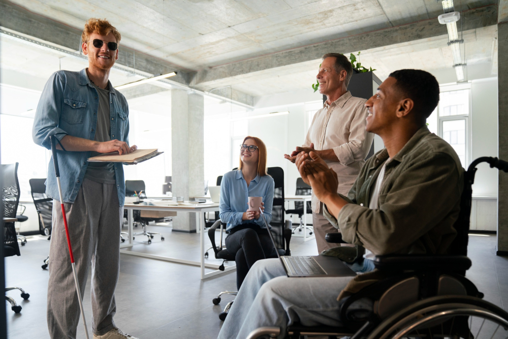 Tecnología para la Vida TXLV - Micro Magazine de FUNTESO, Fundación Tecnología Social imagen en la que destacan 4 personas en una oficina de trabajo, una de ellas sentada en una silla, otra en una silla de ruedas, y dos de pie, siendo una de ellas un hombre ciego con gafas y bastón. Todos están en un ambiente distendido, sonrientes